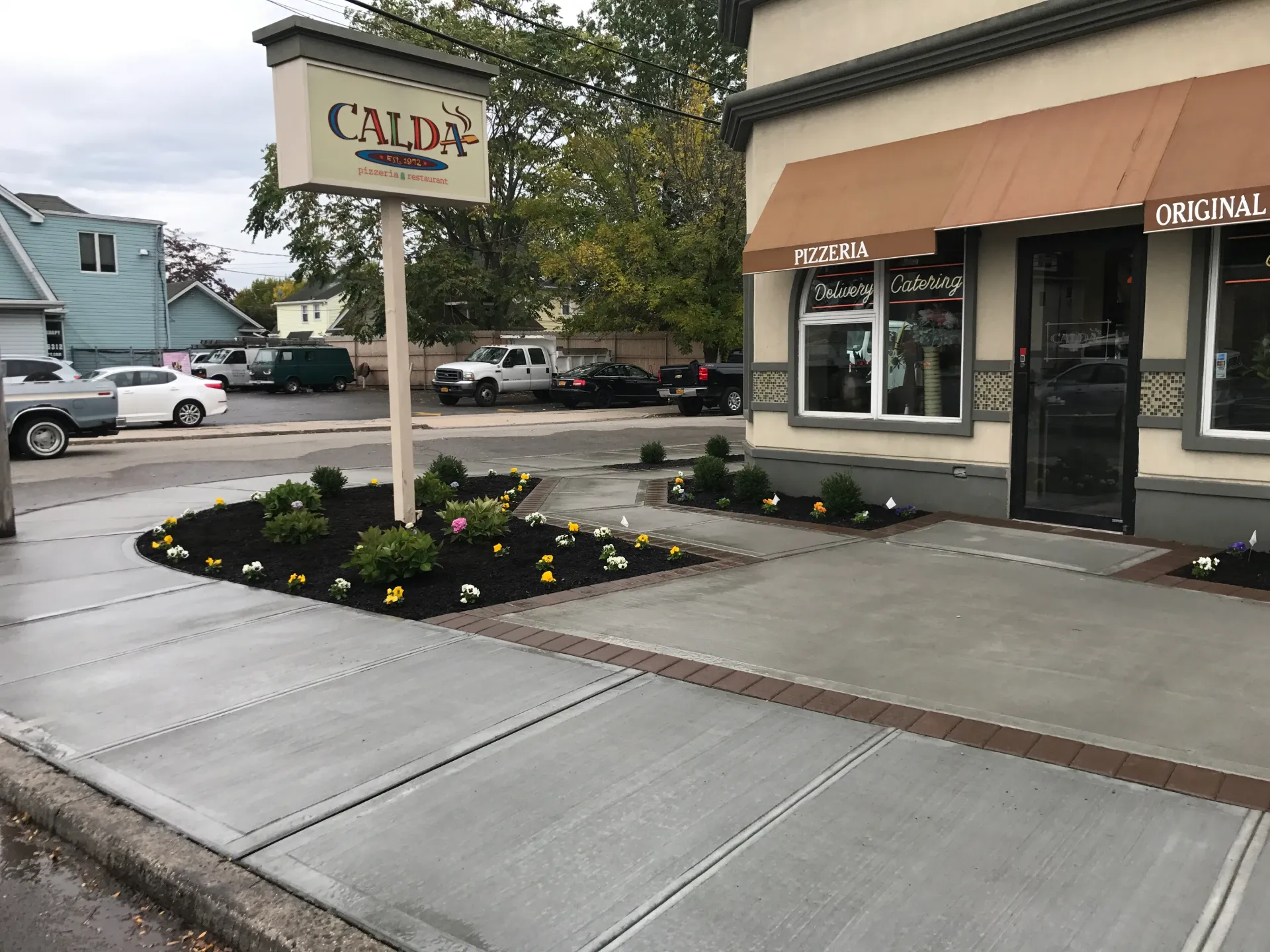 Exterior of Calda's restaurant with a tan awning, sign, and sidewalk planters with flowers. Parking lot in background.