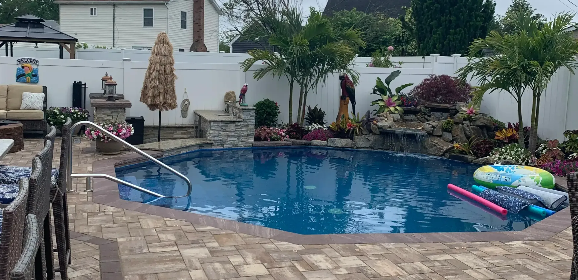 A large swimming pool is surrounded by a brick patio and a white fence.