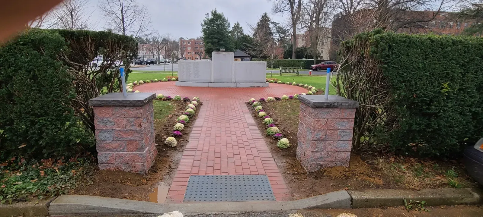 Brick path leads to a monument, bordered by hedges and small flowerbeds, on a cloudy day.