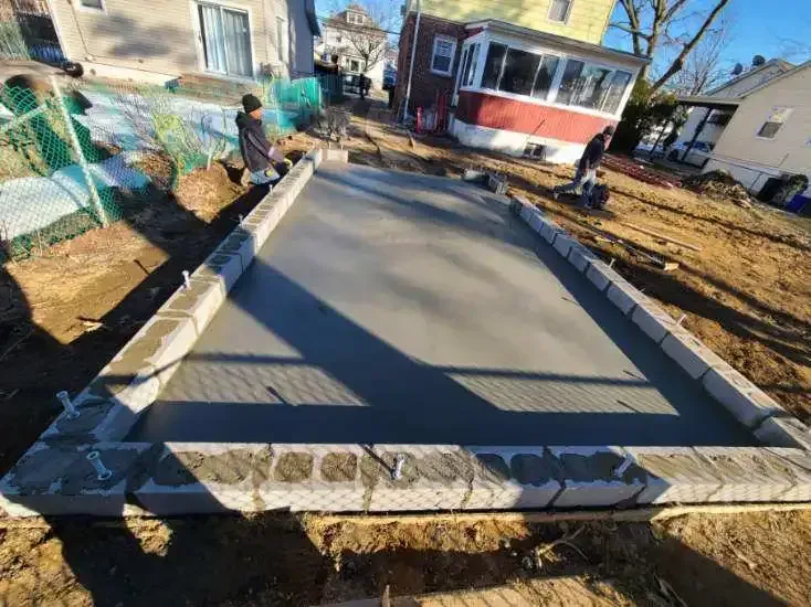 Concrete slab construction in a backyard, bordered by concrete blocks. Two people working on the wet concrete on a sunny day.