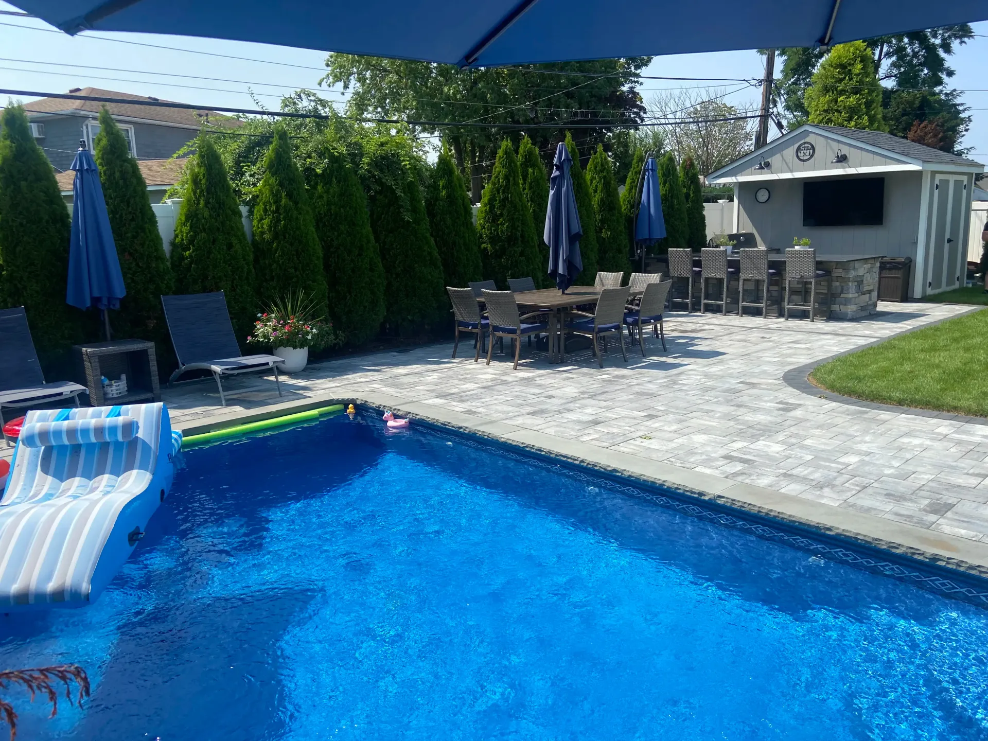 Backyard with pool, patio, dining set, bar area, and green hedges.