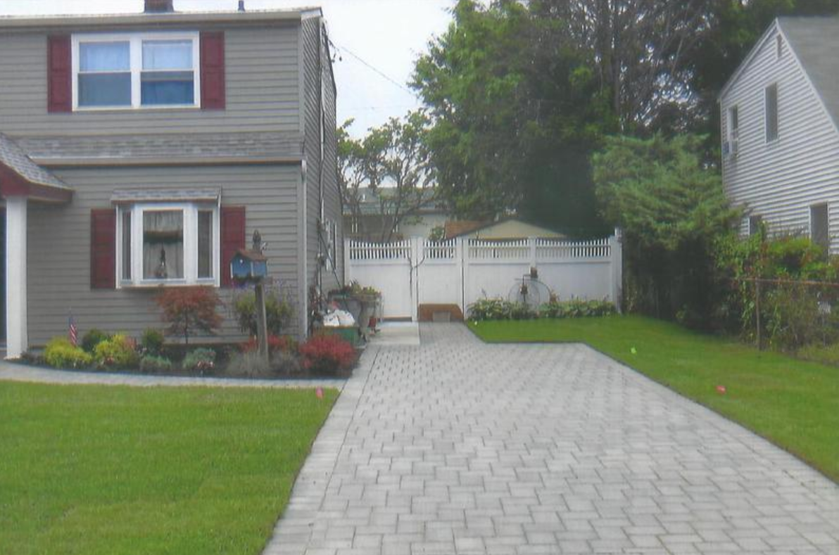 Gray house with a brick driveway and white picket fence, green lawn, and red shutters.