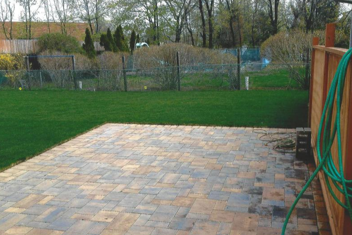 Brick patio next to a lush green lawn, wooden fence with a green hose, trees in the background.