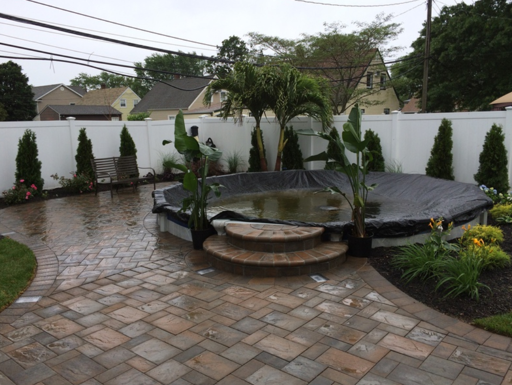Backyard with a round pool, brick patio, and white fence, with lush greenery and a covered pool.