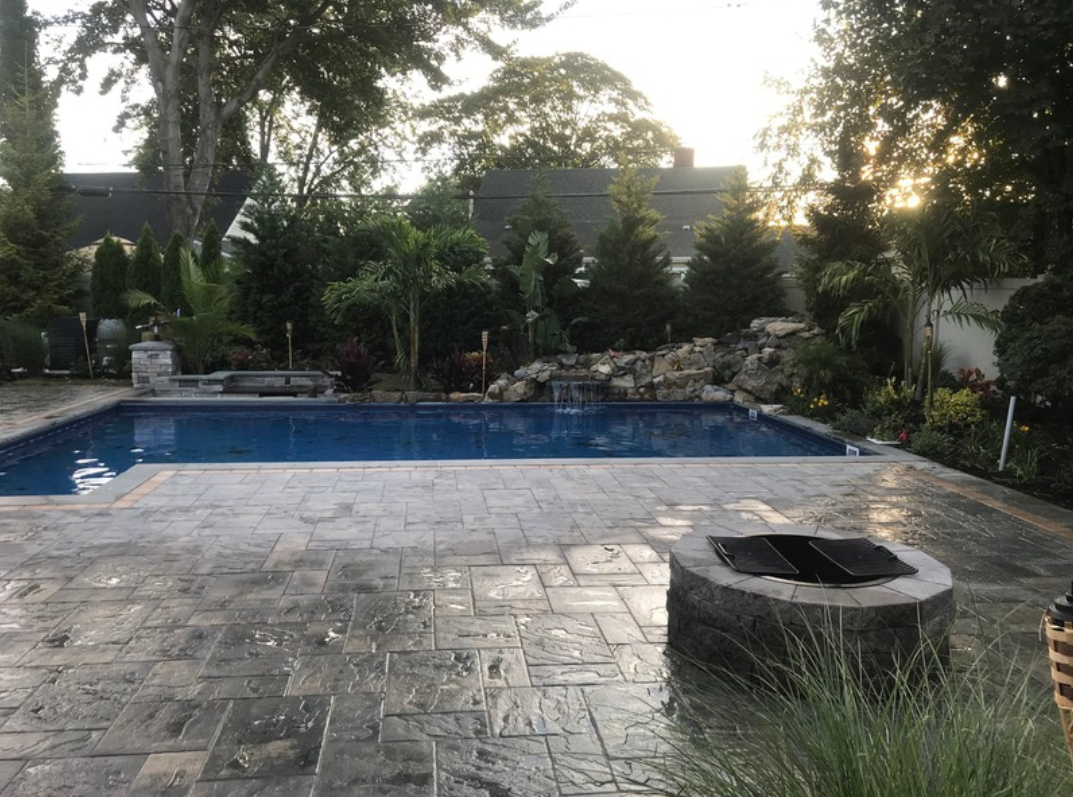 Backyard pool with stone patio, fire pit, and lush greenery.