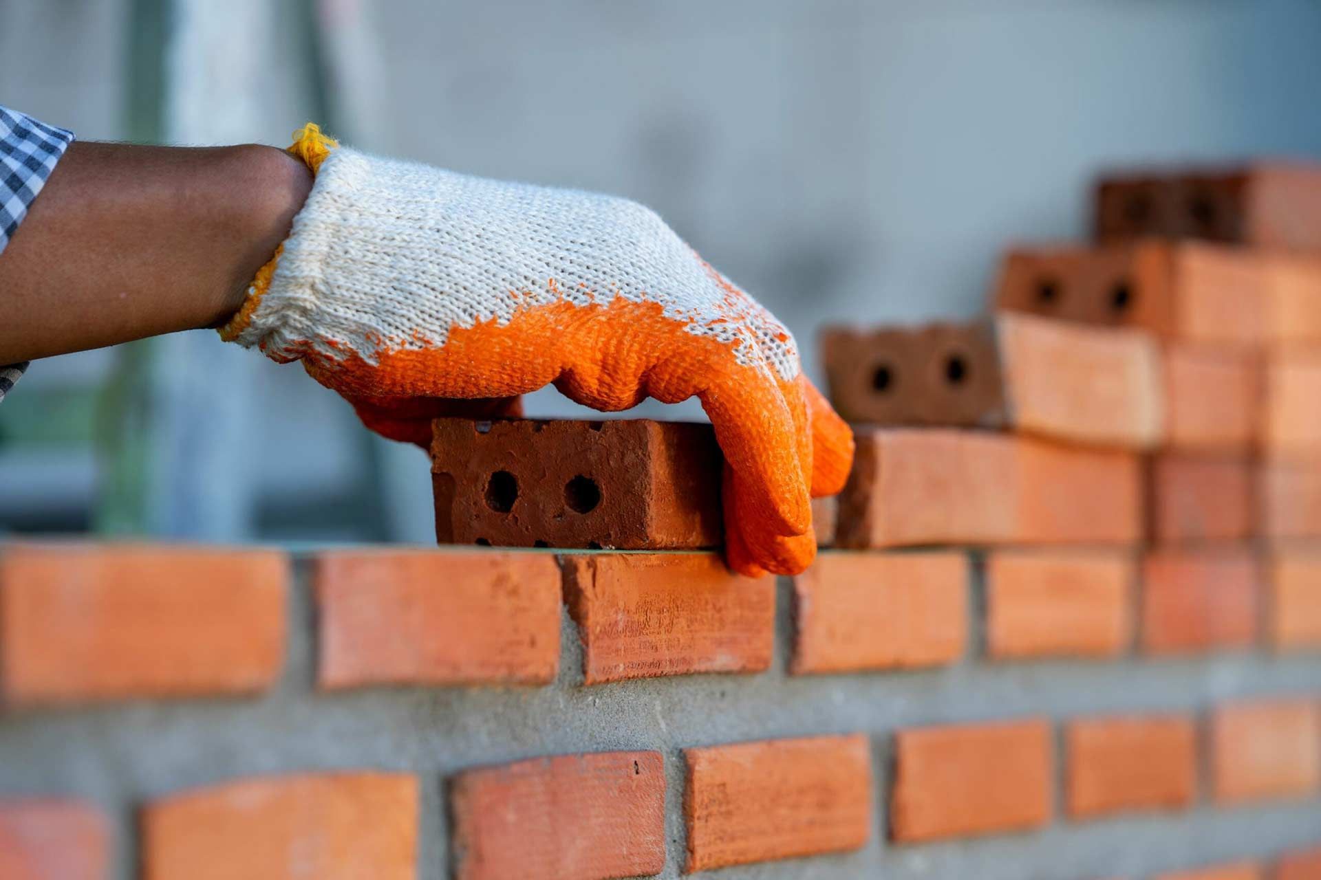 Hand in orange glove laying a brick on a brick wall being built.