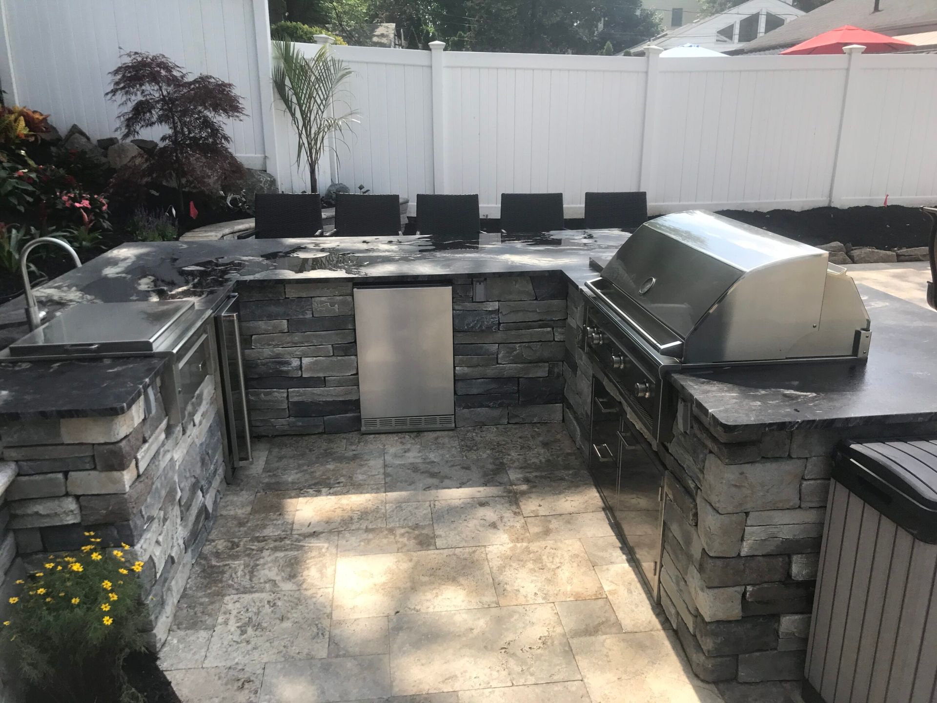 Outdoor kitchen with stone facade and built-in grill, sink, and refrigerator, in a backyard setting.