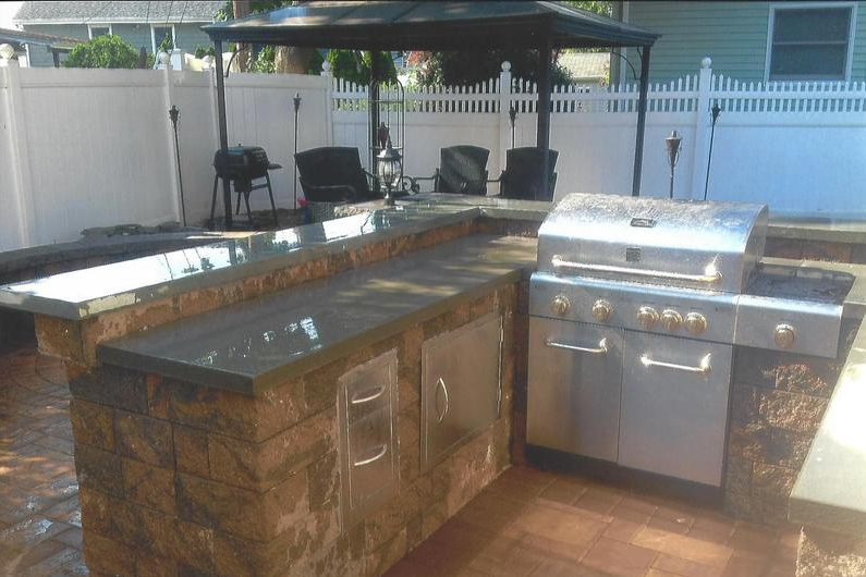 Outdoor kitchen with a built-in grill, counter, and seating under a gazebo, next to a white fence.