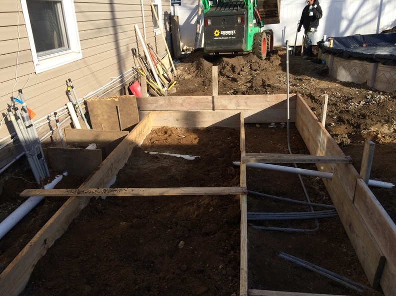 Wooden forms for a concrete project, dirt inside. A Bobcat and worker are in the background.