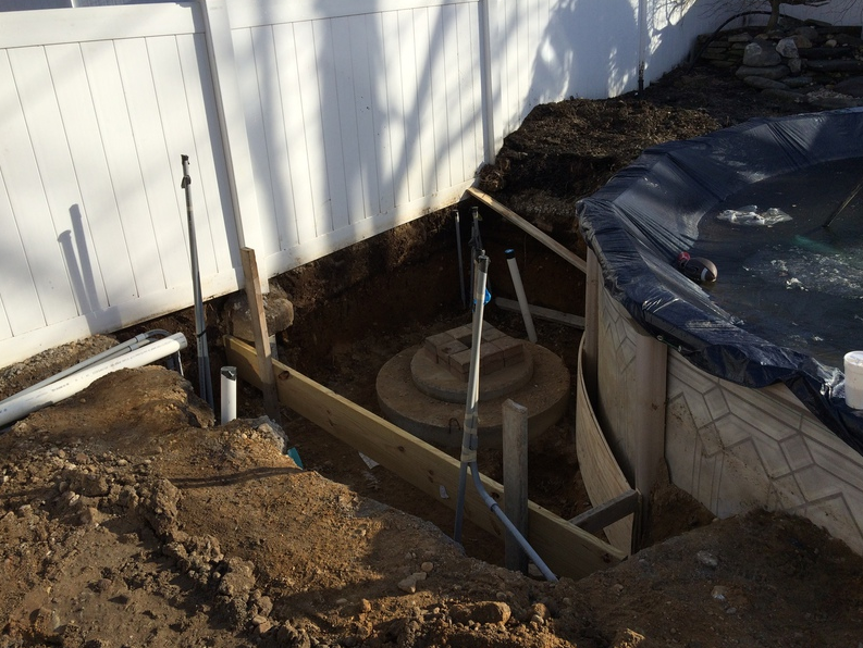 Construction site with a pool, wooden frame, pipes, and earth against a white fence.
