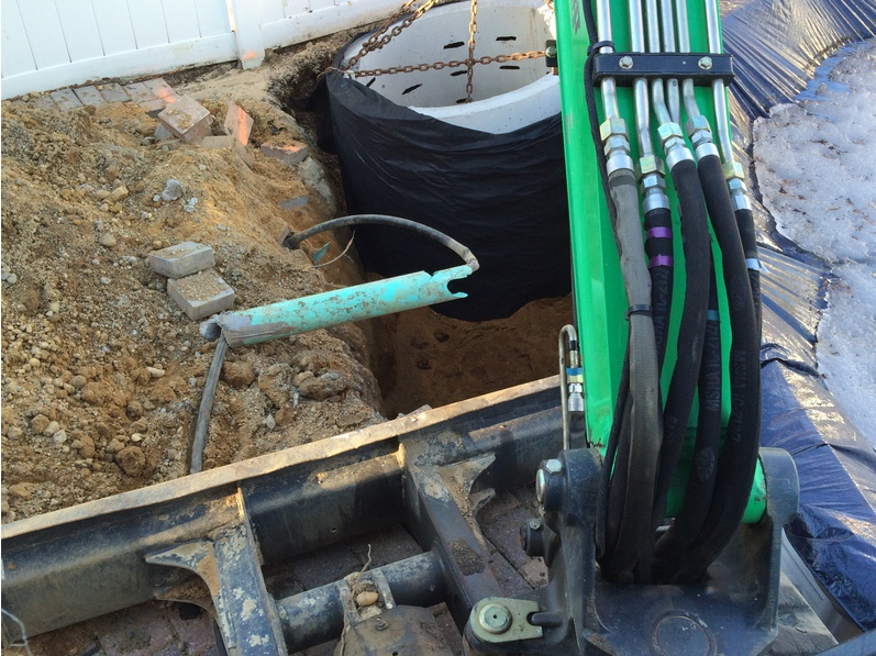 An excavator digging a hole next to a white fence; a large black container is visible in the hole.