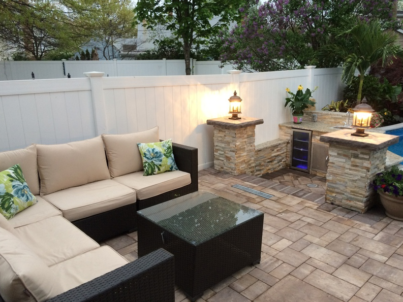 Outdoor patio with sectional sofa, stone accents, and a small kitchen area, all enclosed by a white fence.