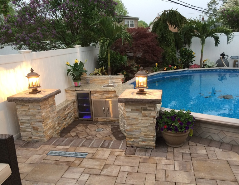 Outdoor kitchen with a pool in the background; stone counters, lights, and a cooler.