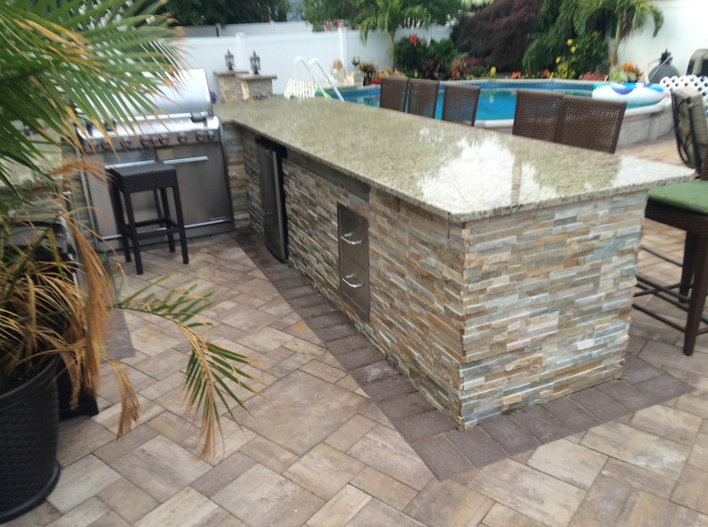 Outdoor kitchen with granite countertop, stone facade, grill, refrigerator, and bar seating.