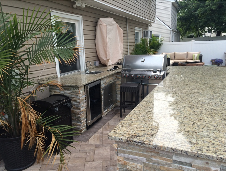 Outdoor kitchen with a grill, sink, and counter. Granite countertops, brick facade, and palm tree.