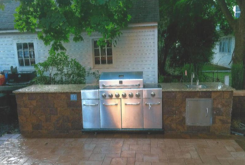 Stainless steel outdoor grill and countertop with a brick facade, next to a house.