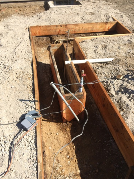 Wooden structure in a dirt trench, with wires and pipes, likely a construction or testing setup outdoors.