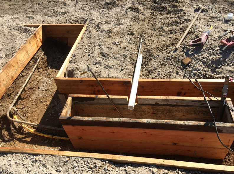Wooden forms for concrete structure, with pipes and tools on dirt ground outside.