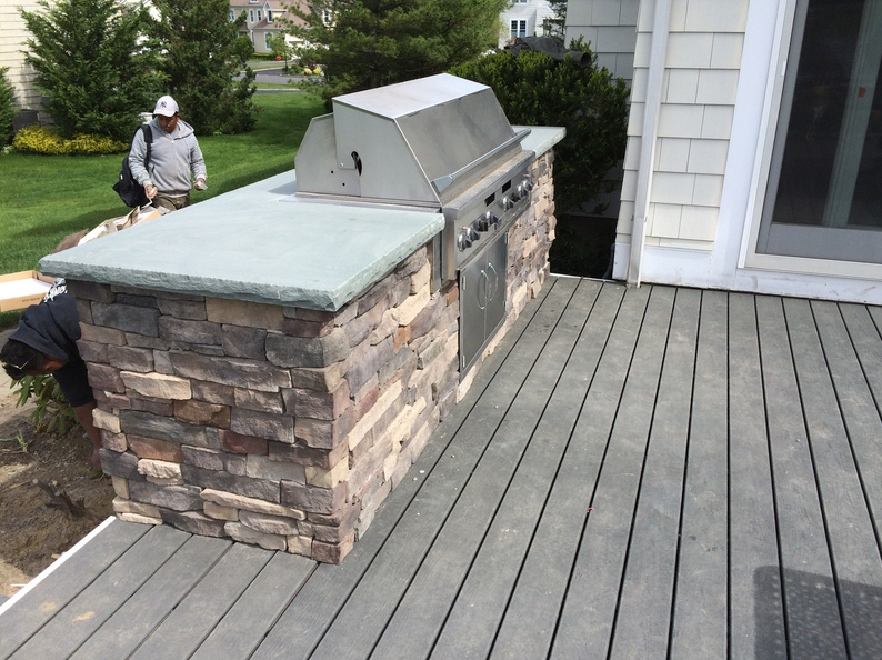 Stone outdoor kitchen with a grill on a wooden deck. A person is working in the yard.
