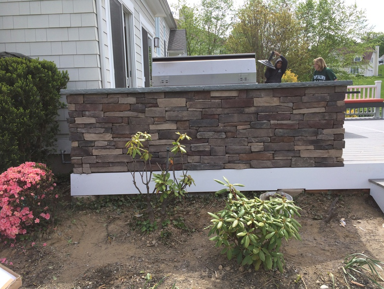 Stone outdoor kitchen with grill, shrubs, two people, and a white house exterior.