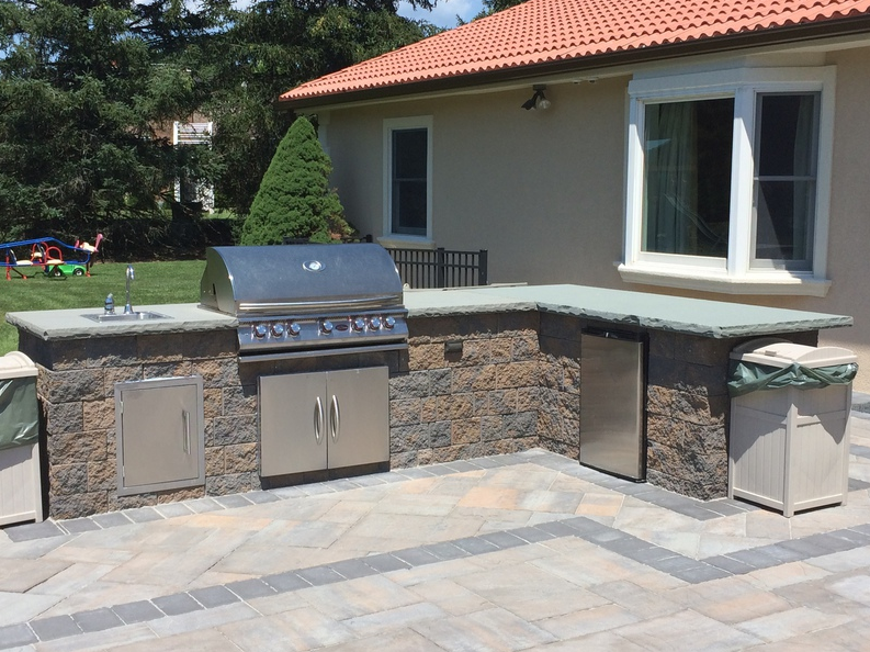 Outdoor kitchen with grill, sink, fridge, and stone countertop; tan and grey patio.