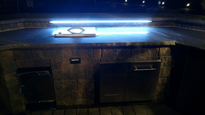 Outdoor kitchen with dark countertop, brick facade, stainless steel cabinet, and overhead LED lighting.