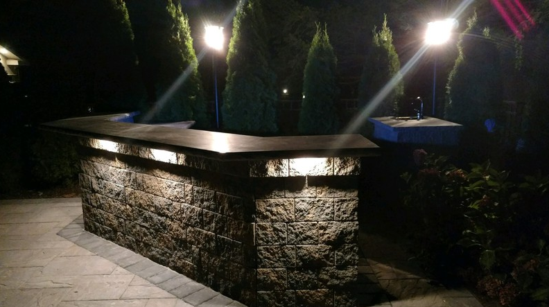 Nighttime outdoor bar with stone facade and illuminated countertop, surrounded by trees and spotlights.