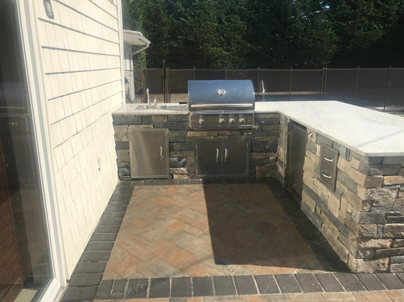 Outdoor kitchen with stone surround, grill, and built-in appliances. Brown and beige stone, gray counter, brick floor.