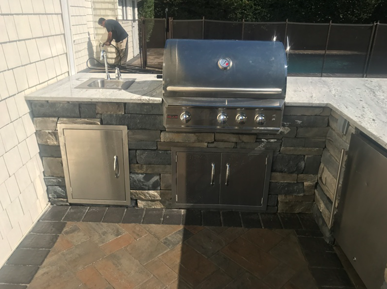 Outdoor kitchen with grill, sink, stone surround, and stainless steel doors. A person is near the sink.