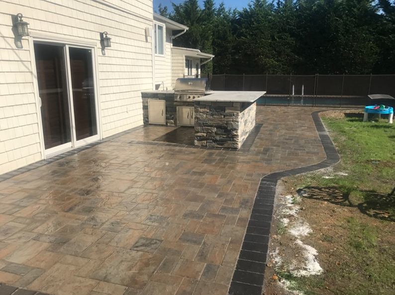 Backyard patio with a stone-covered outdoor kitchen and a paved pathway, bordered by grass and a fence.
