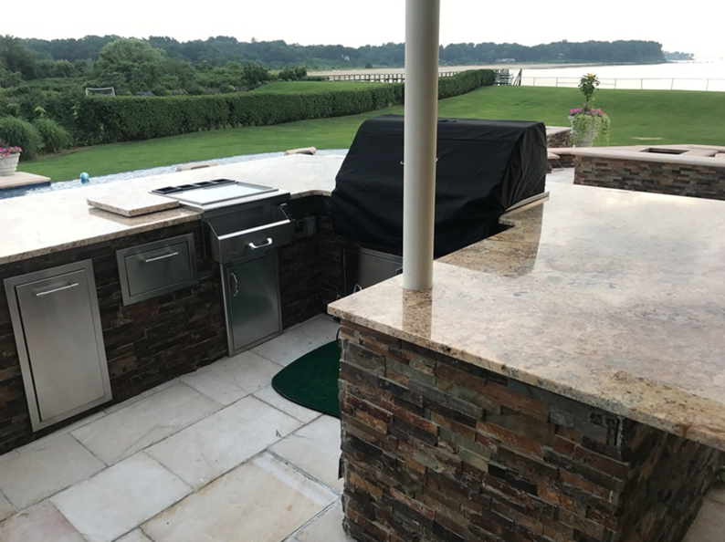 Outdoor kitchen with stone and granite counters, grill, and storage. Overlooks a lawn and water.