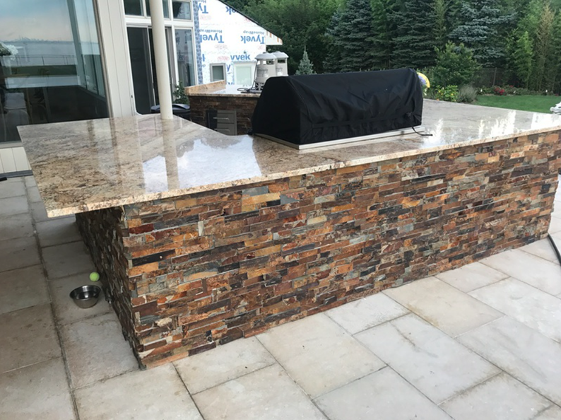 Outdoor kitchen island with stone facade, granite countertop, and grill under a cover.
