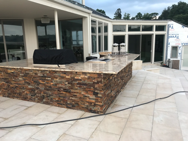Outdoor kitchen with stone facade, granite countertop, and grill, next to a modern house on a patio.