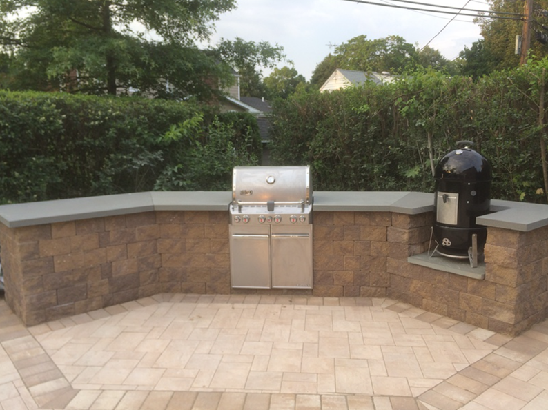 Outdoor kitchen with built-in grill and smoker on a brick patio, surrounded by stone walls and greenery.