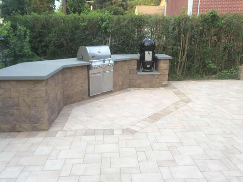Outdoor kitchen with grill and smoker on a stone patio, surrounded by greenery.