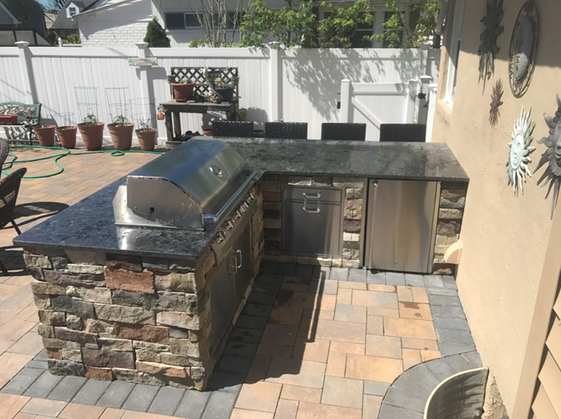 Outdoor kitchen with granite countertop, grill, and fridge, set on brick patio.