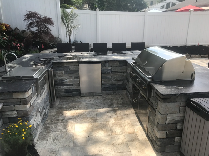 Outdoor kitchen with stone facade, stainless steel grill, sink, and built-in refrigerator, on a tiled patio.