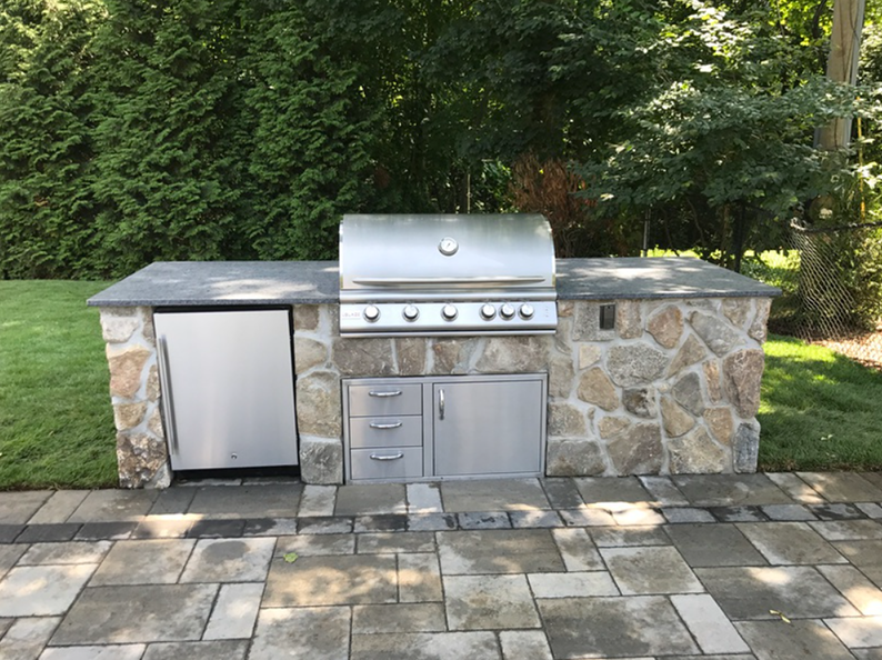 Outdoor stone grill with a refrigerator and storage, set on a patio with a grassy background.