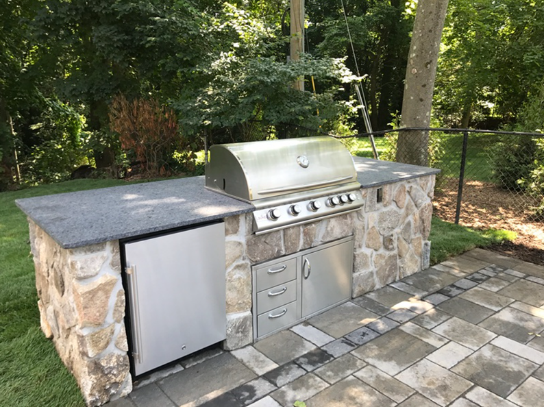 Outdoor kitchen with stone facade, stainless steel grill, refrigerator, drawers, and countertop on a patio.