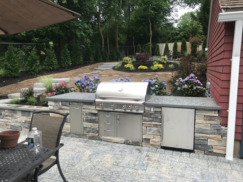 Outdoor kitchen with grill and built-in refrigerator on a stone patio. Landscaped backyard with flowers.