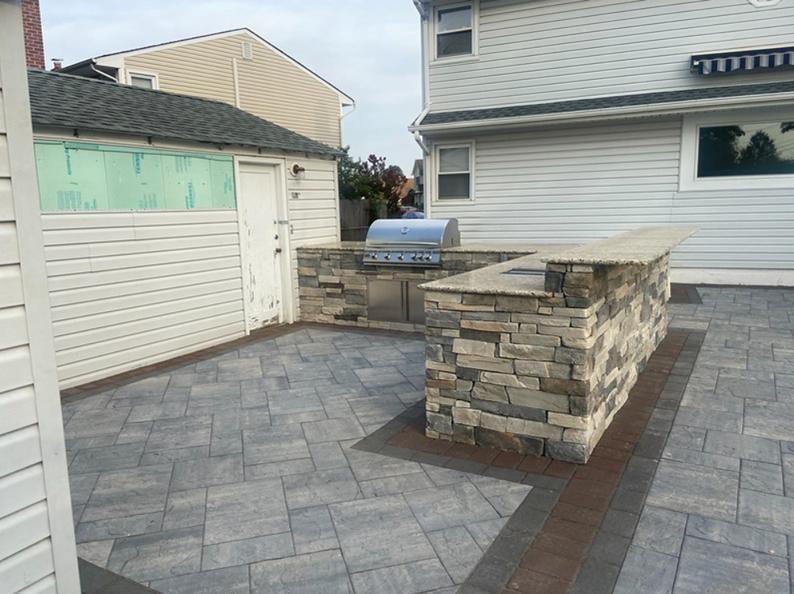 Outdoor kitchen on a paved patio with a grill and bar made of stone, next to a white building.