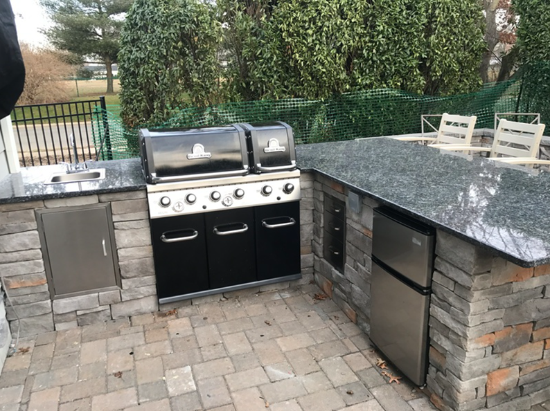 Outdoor kitchen with a grill, sink, fridge, and granite countertops on a stone patio.