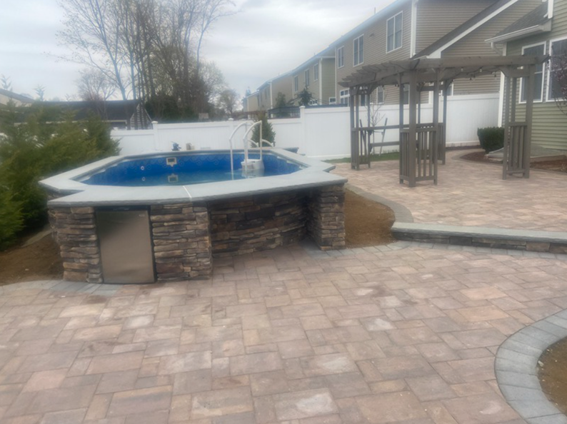 Backyard with a small pool, patio, and pergola. Beige pavers, stone pool surround, and a stainless steel door.