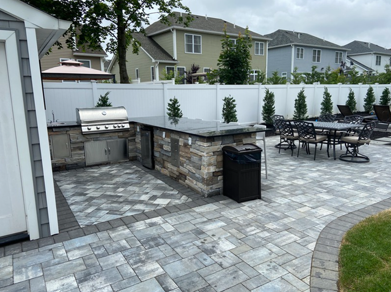 Outdoor kitchen on a paved patio, with grill, bar, and dining set; residential setting.