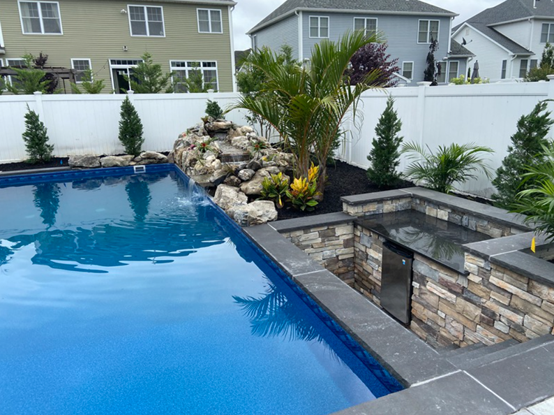 Pool with waterfall, surrounded by landscaping and stone accents. Blue water, white fence.