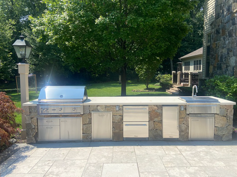 Outdoor stone-clad kitchen with stainless steel grill, storage, and sink on a patio.