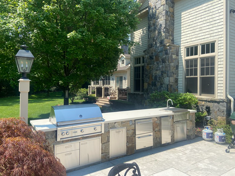 Outdoor kitchen with stainless steel appliances, built-in grill, and stone facade next to a house.