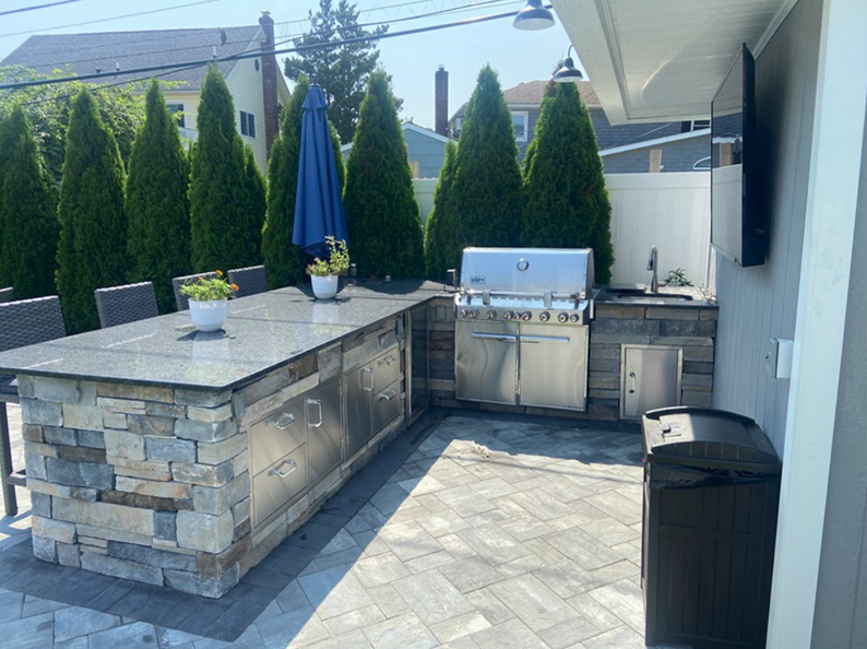 Outdoor kitchen with a grill, counter, and seating. Stone facade, blue umbrella, and tall green trees in the background.