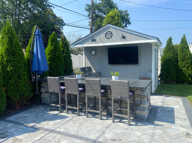 Outdoor kitchen with stone bar, gray stools, grill, TV, and blue umbrella.