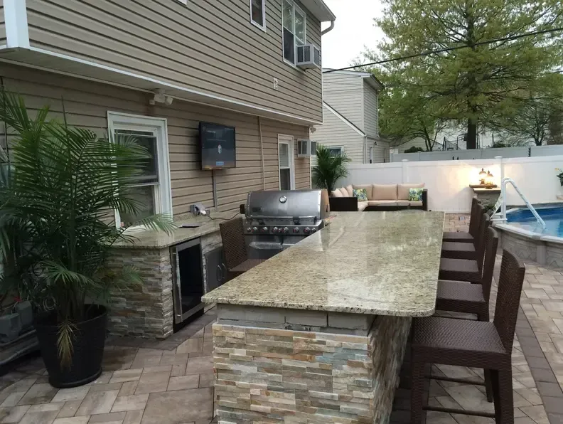 Outdoor kitchen with granite countertop and built-in grill, near a pool and seating area.